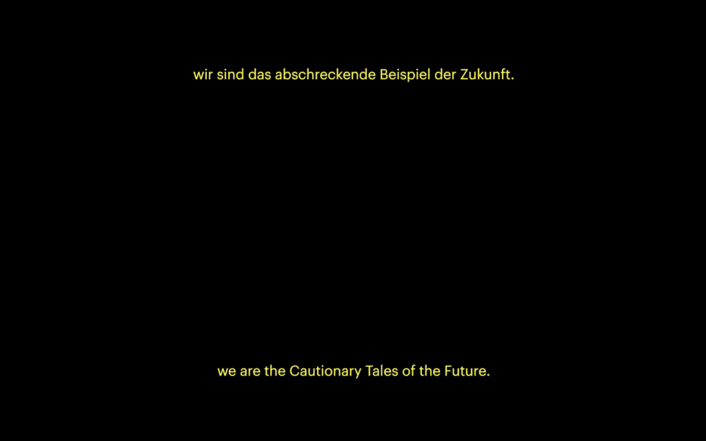 A still of a black video screen with subtitles in yellow sans serif text at the top and bottom. The top layer of subtitles are in German. The bottom layer reads "we are the Cautionary Tales of the Future".