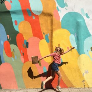 a young blonde woman jumping in the air against the backdrop of a multi-colour painted wall
