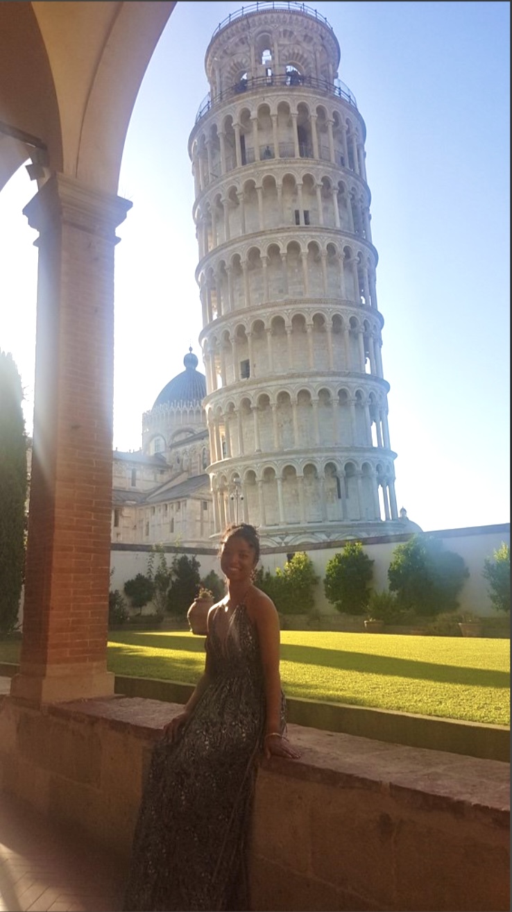 Lydia, a young woman, sitting on a short brick wall with the leaning tower of Pisa in the backdrop.