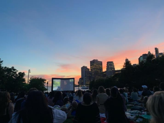 crowd of people sitting on ground outside in a park watching a movie on big screen with orange sunset and city skyline behind
