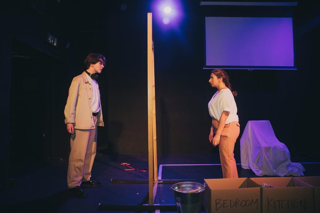 Liv, wearing a beige shirt and trousers, stands stage right looking through a large wooden frame into the eyes of another performer, also wearing beige