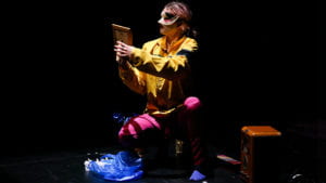 A figure wearing a yellow top, pink trousers and a decorative eye mask crouches down looking at a picture in a frame